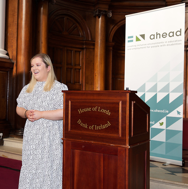 Tessa Lyons standing beside a podium at the WAM Leaders Award in 2022. Tessa has shoulder length blonde hair and is wearing a white dress with black spots.