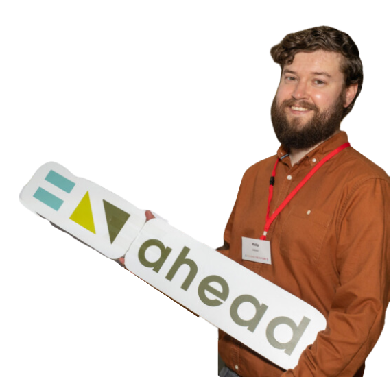 Philip has brown hair and a beard and wearing a brown shirt holding the AHEAD logo 