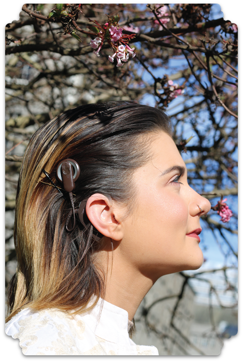 A colour photograph taken on a bright day of a young adult female. She faces to the right hand side and in doing so reveals a cochlear implant in her long straight brown hair. In the background we see bare branches of a tree with just small pink flowers on the tips.